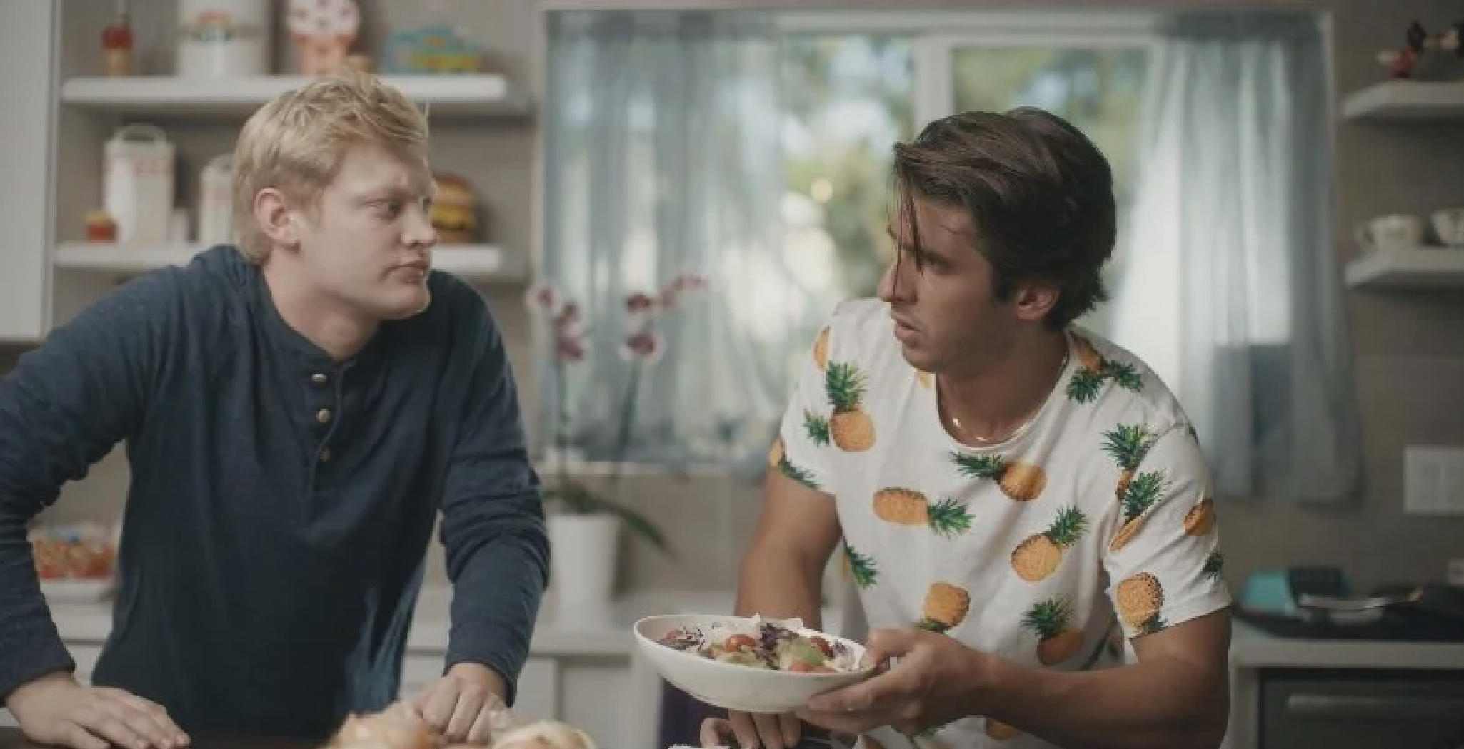 Two men are engaged in a conversation in a kitchen. One man has blonde hair, is wearing a dark blue long-sleeve shirt. The other man Paul Santoli has dark hair, is wearing a white t-shirt with pineapple print, and is holding a bowl of salad.