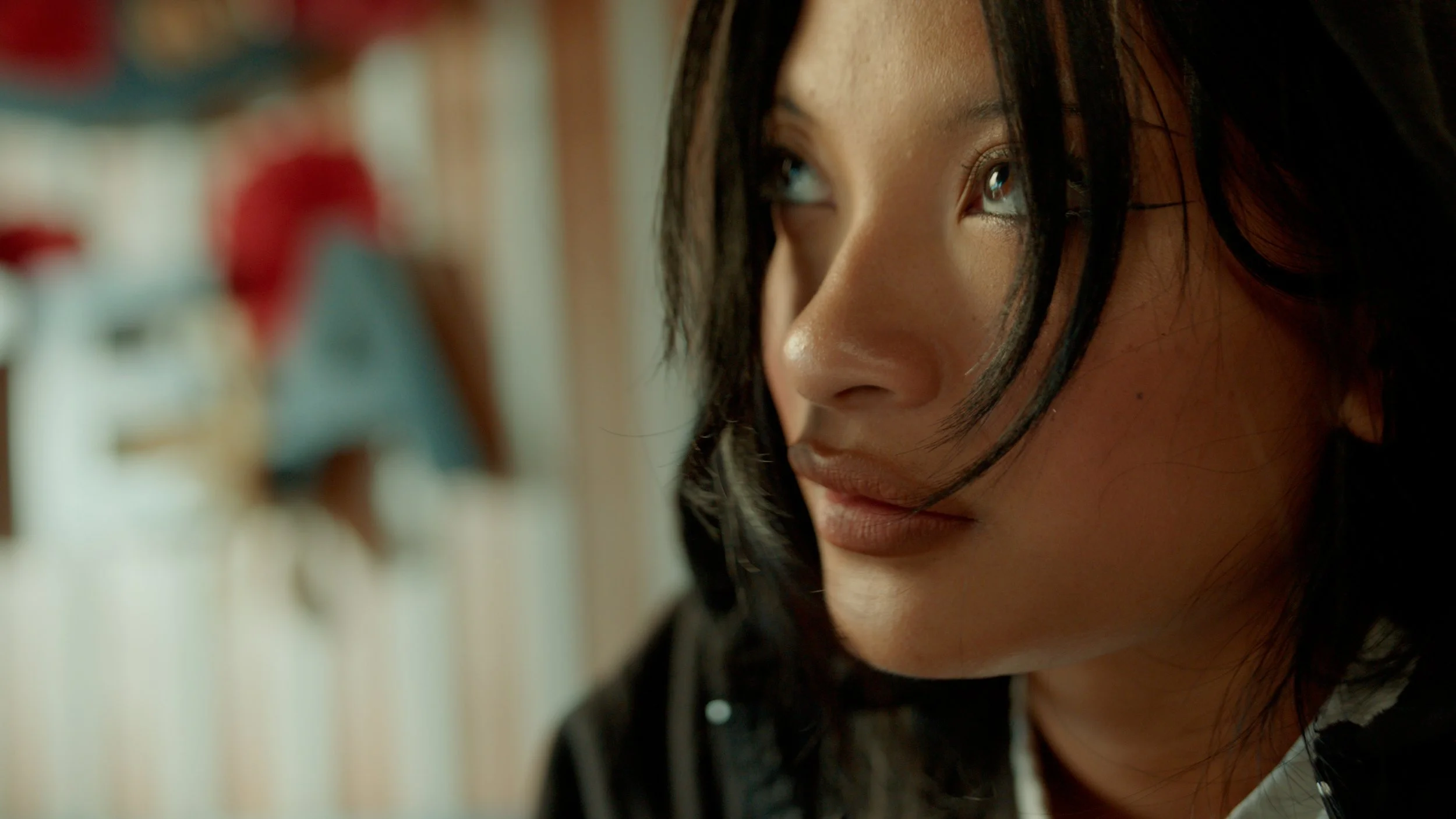 Close-up of a young woman with black hair, light skin, and blue eyes, looking to the side with a thoughtful expression. The background is blurred with colorful objects.