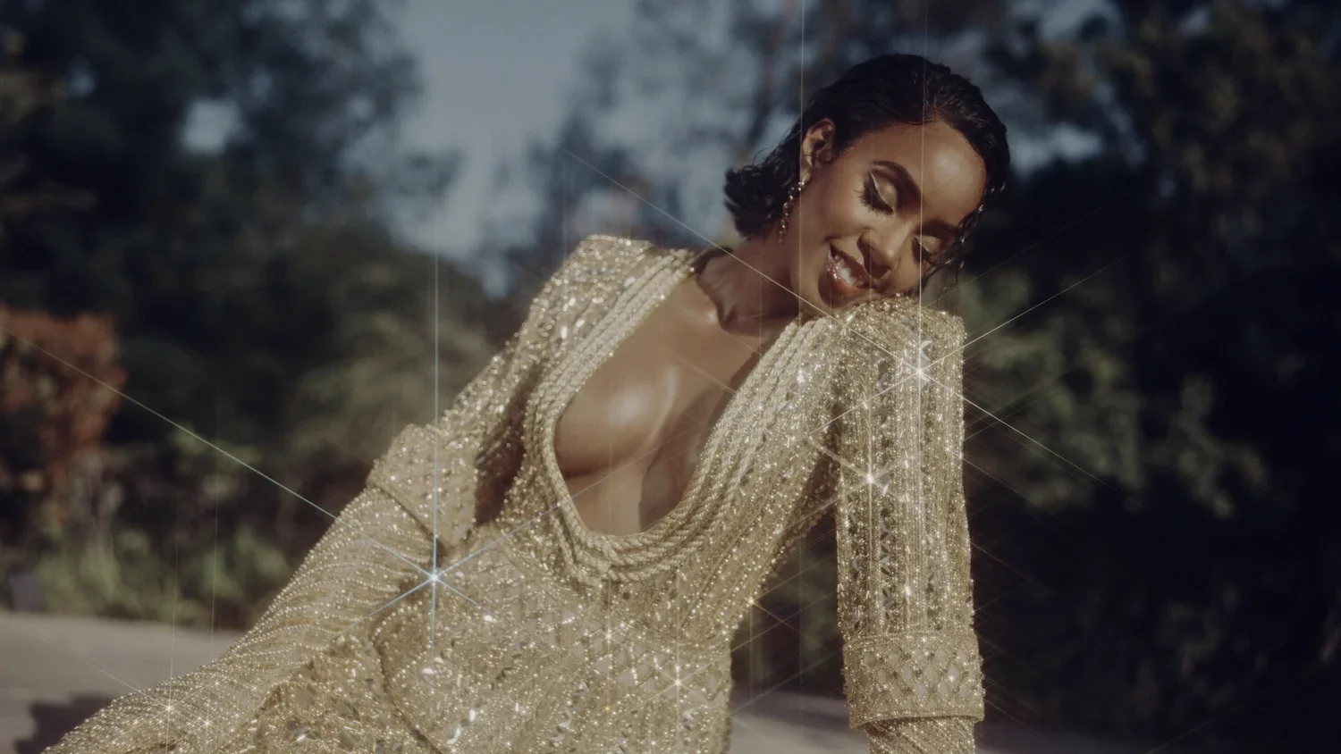 Woman in a gold, sequined dress outdoors, Kelly Rowland, Joshua Libertine smiling and looking down with eyes closed.