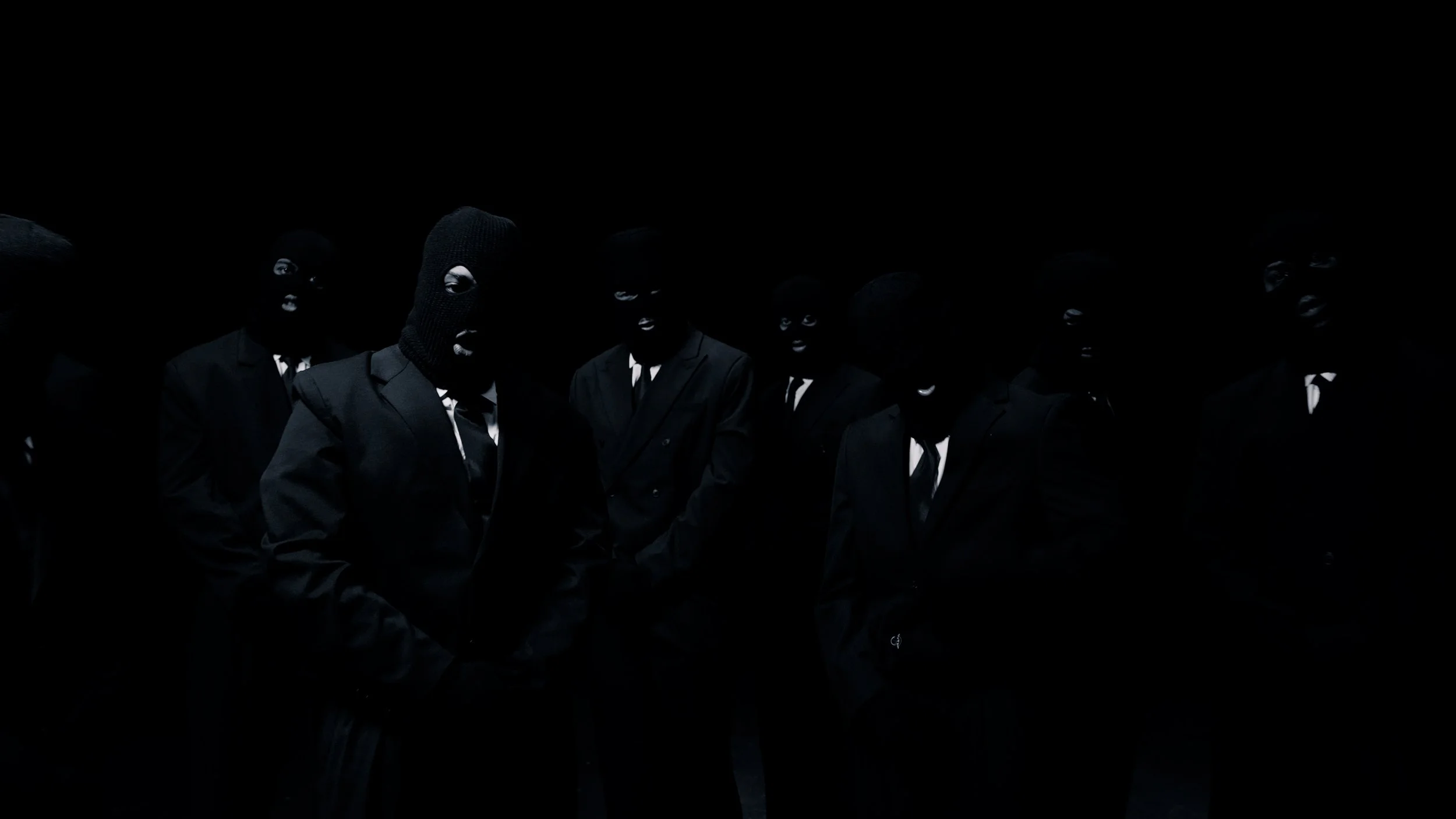 Group of people dressed in black suits wearing black ski masks, standing in a dark setting.