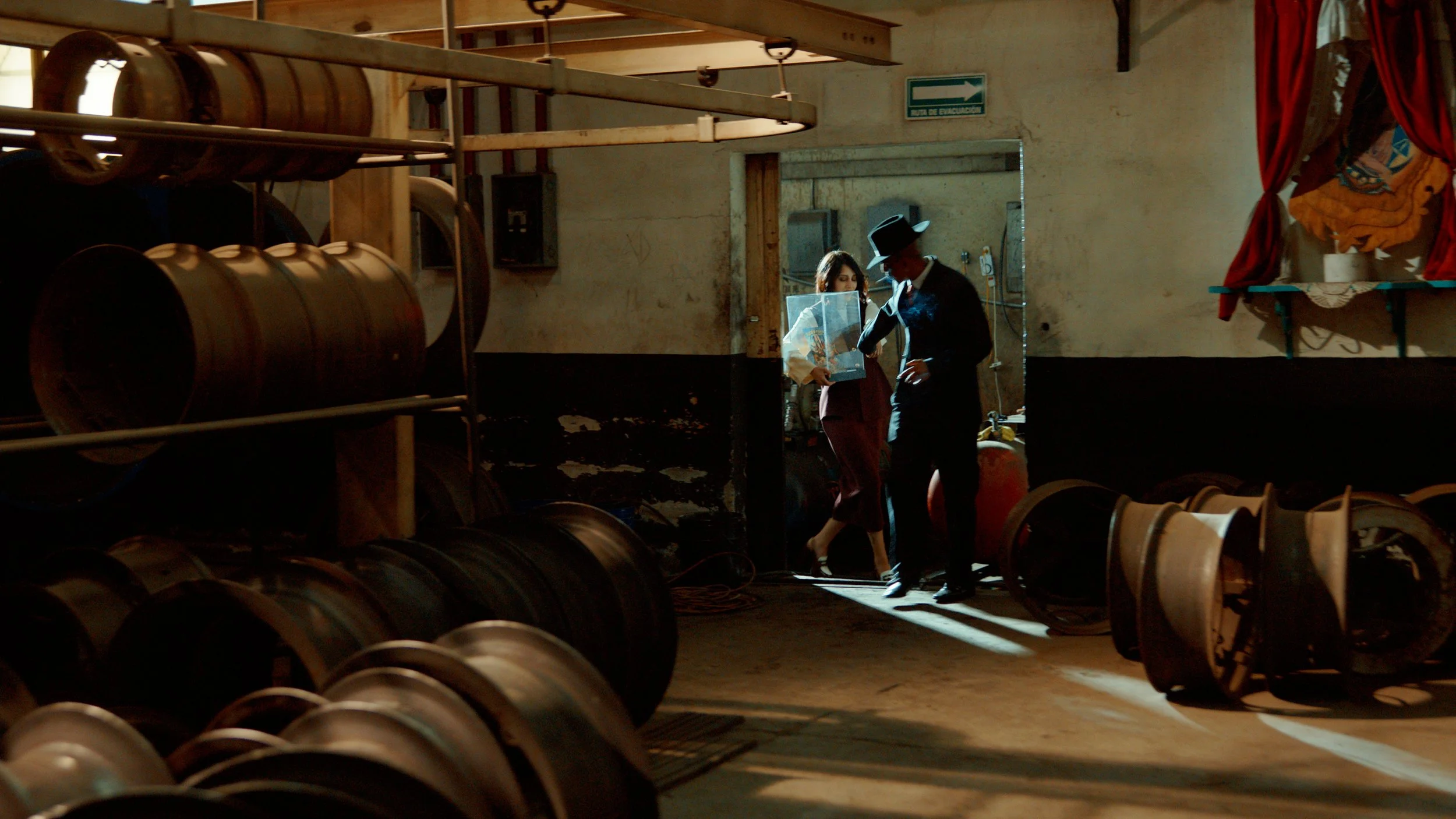 Two people in vintage clothing standing in a rustic room with shelves of large industrial lights or metal barrels, engaged in conversation.