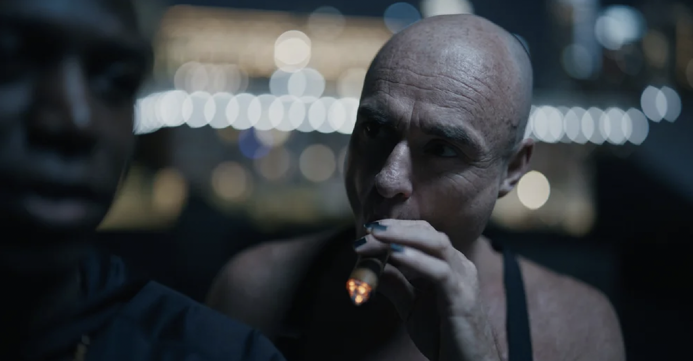 A scene with two individuals, one in focus and one blurred, at night with city lights in the background. The person in focus is a woman with a shaved head smoking a cigarette or cigar.