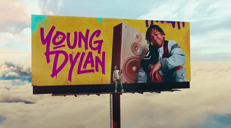 Billboard advertisement for Young Dylan featuring a young boy standing in front of it, with a large image of a smiling young boy and stylized speaker graphics on the billboard.