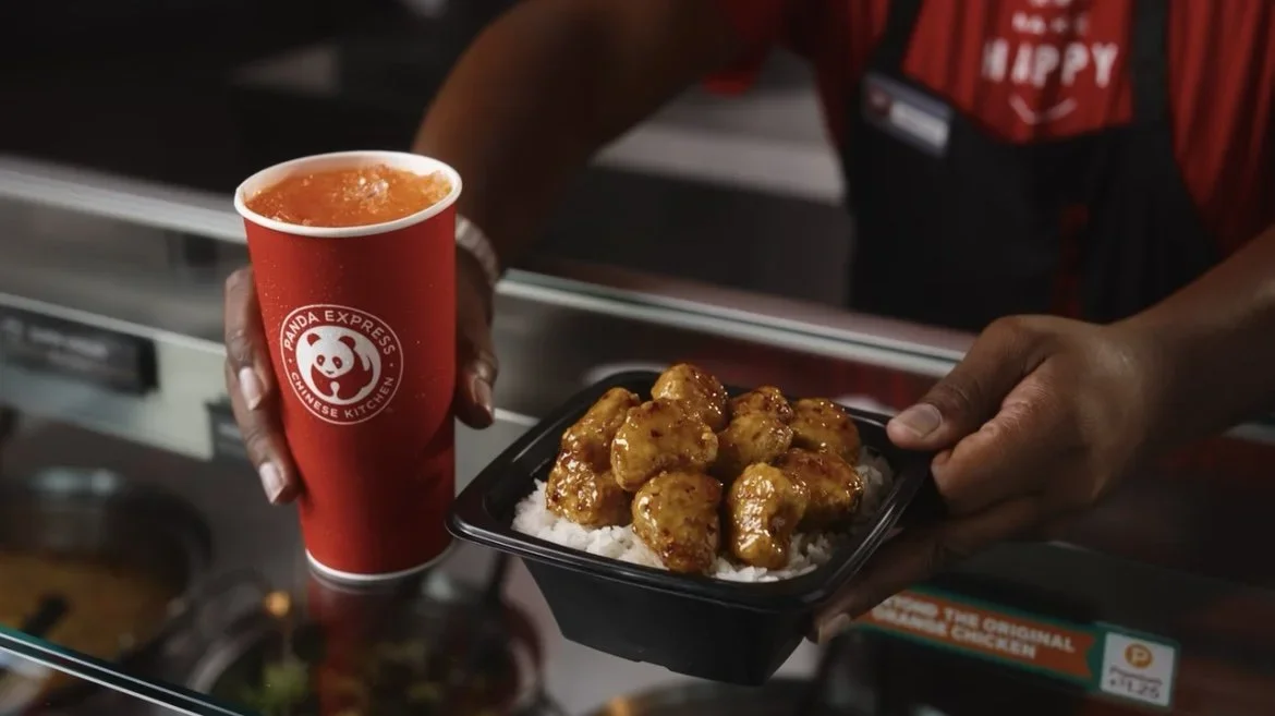 A person holding a red Panda Express cup with orange-colored beverage and a black tray with orange chicken served over white rice.