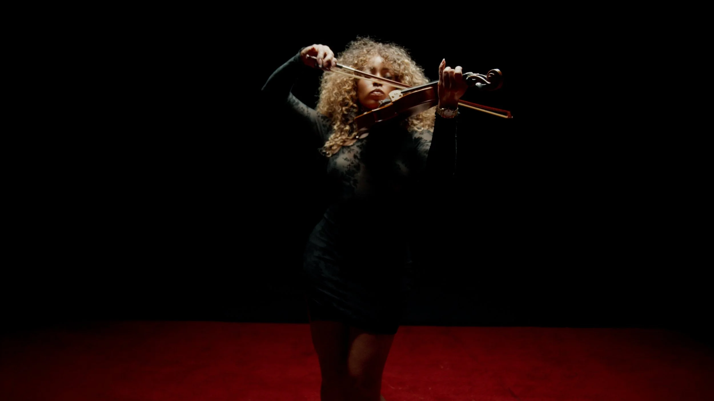 Suzette James Woman with curly blonde hair playing violin on a dark stage, illuminated by a spotlight, with a red carpeted floor.
