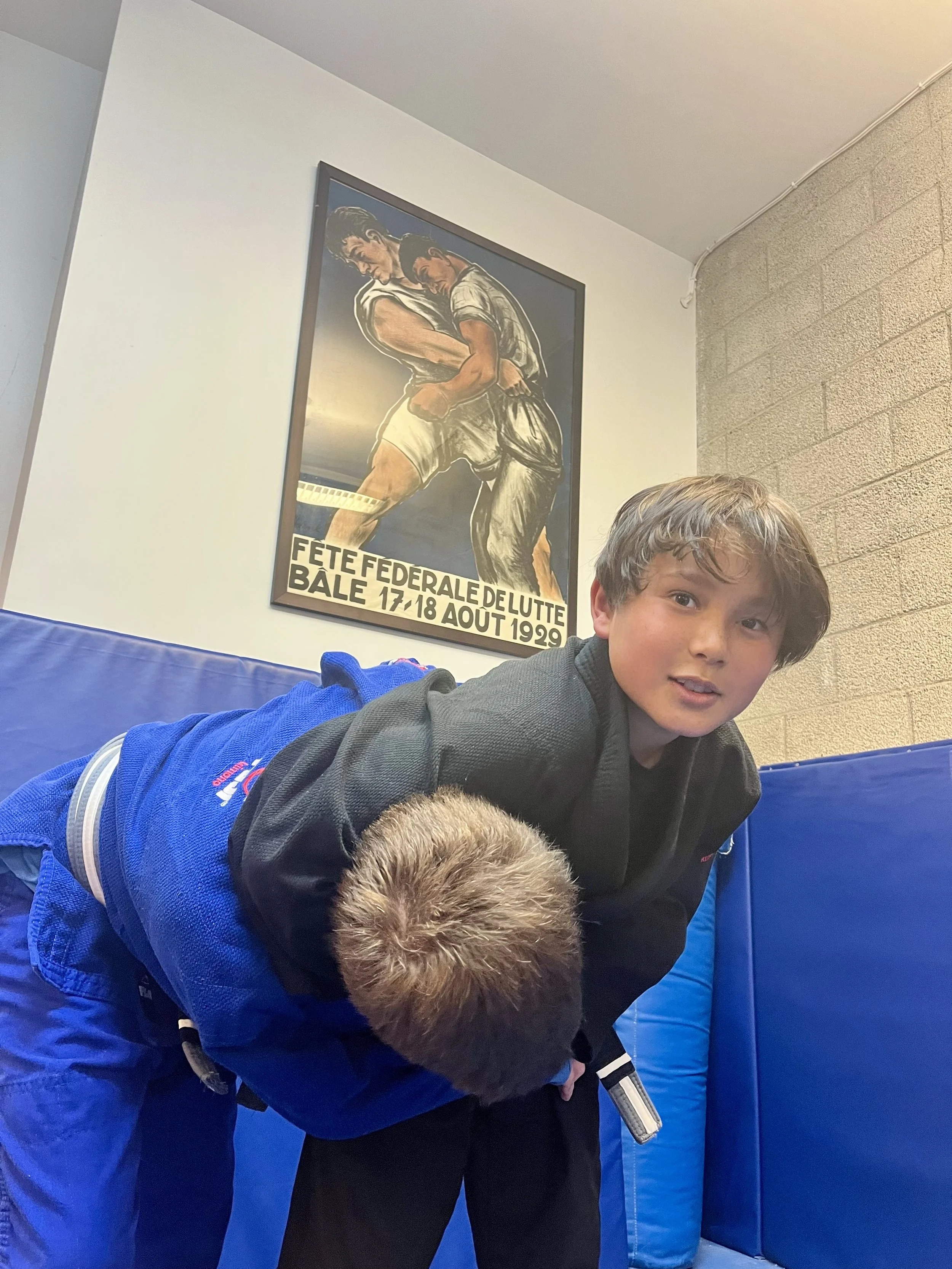 Young boy in a black jiu-jitsu gi uniform practicing jiu-jitsu on a blue padded mat in a gym or training room.