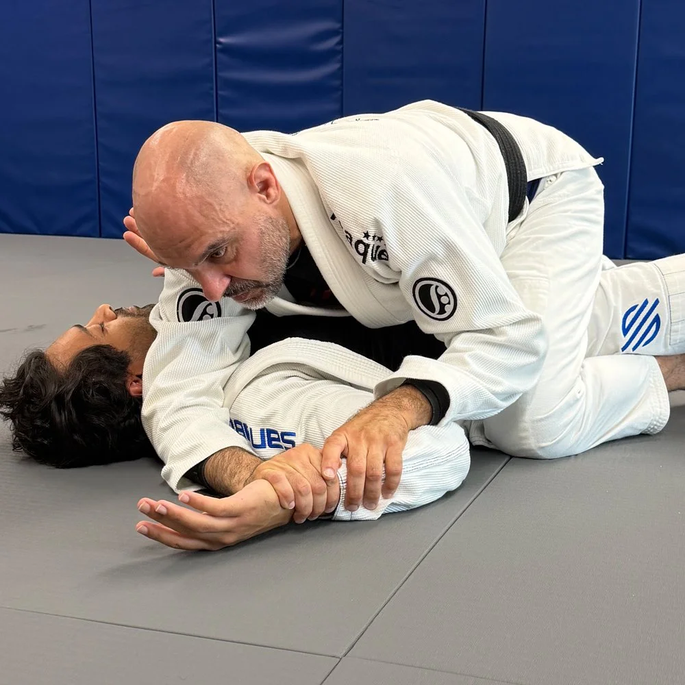 Two Brazilian Jiu-Jitsu practitioners practicing a submission technique on the mat, both wearing white gis. One is a bald man applying a chokehold on an opponent with dark hair.