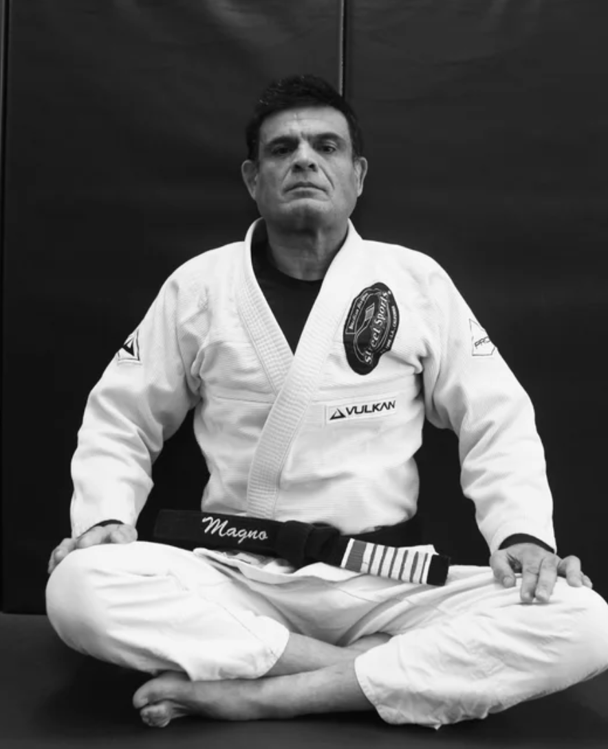 A man in a white Brazilian Jiu-Jitsu gi sitting cross-legged on the mat, looking serious, with a black belt.