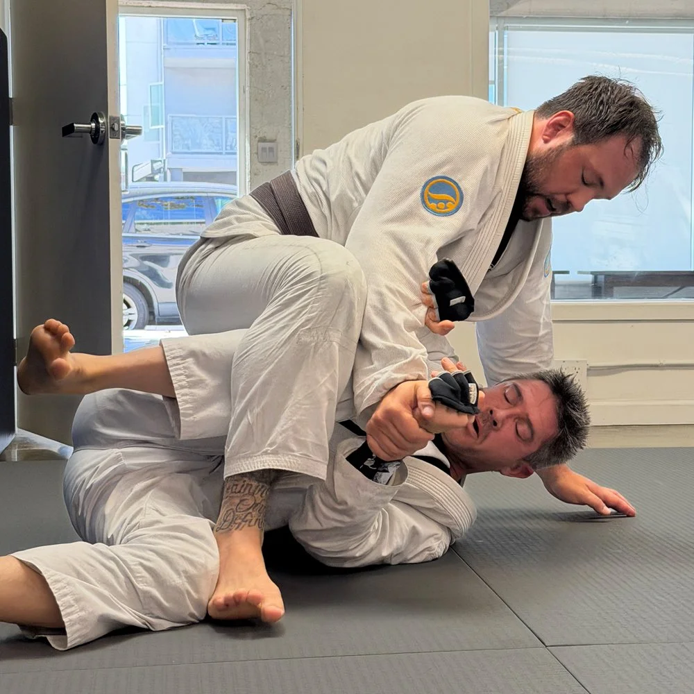Two men practicing Brazilian Jiu-Jitsu on a black mat in a training facility, with one man in a white gi on his back and the other man in a white gi on top, both wearing black gloves. The man on top is pinning the man on the bottom, who has tattoos on his left ankle and is holding a small object in his mouth.