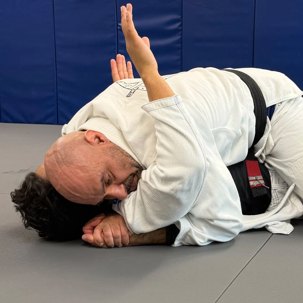 Two people practicing Brazilian Jiu-Jitsu on a mat; one is wearing a white gi and the other a black gi, with the person in black on top.