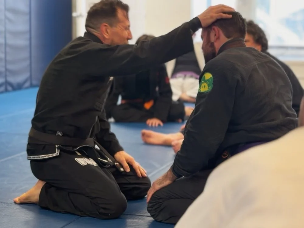 Martial arts instructor showing technique to male student during class, with onlookers in the background.