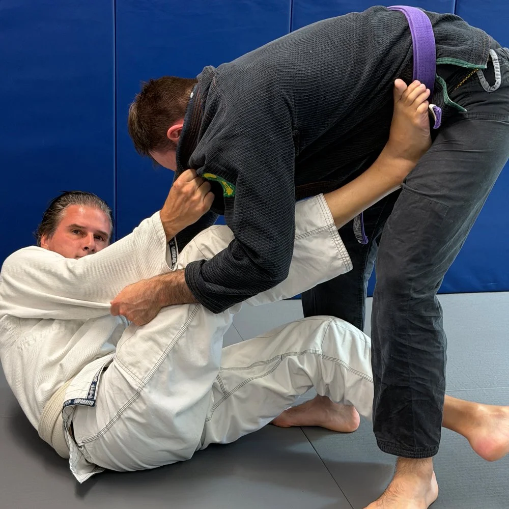 Two men practicing Brazilian Jiu-Jitsu on a mat, with one in a white gi and the other in a black gi, engaged in a grappling technique.