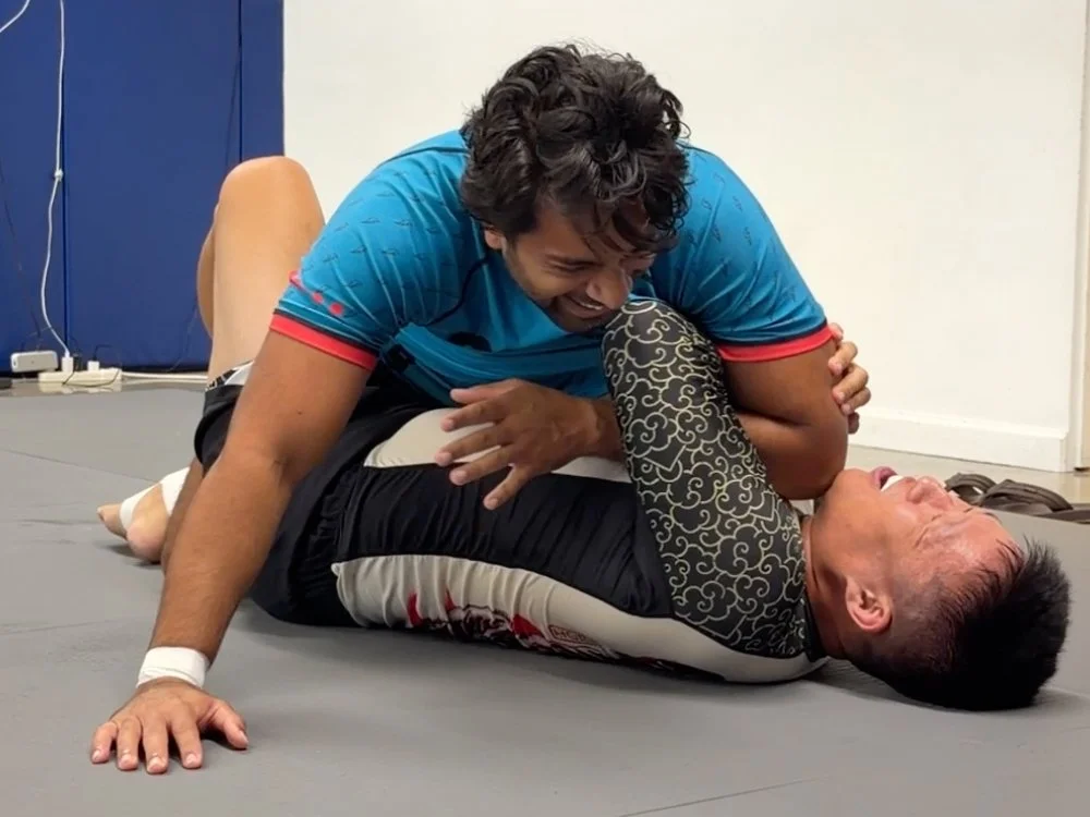 Two men engaged in Brazilian Jiu-Jitsu training on a gray mat, with one man on top and the other on his back, both smiling and having fun.