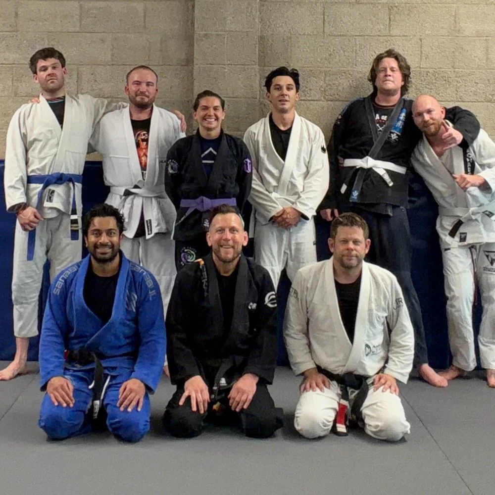 Group of nine people in Brazilian Jiu-Jitsu gi uniforms posing together in a gym.