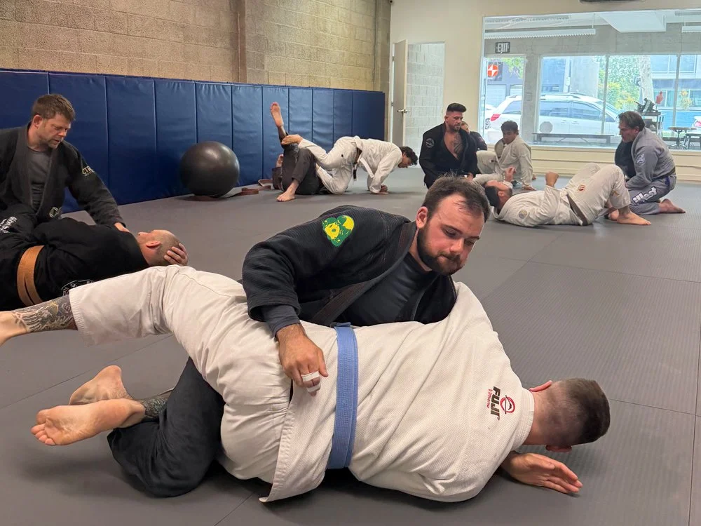 Group of people practicing Brazilian Jiu-Jitsu in a training gym, some on the mat grappling, others stretching or resting in the background.