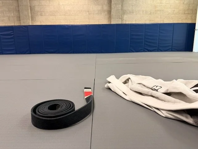 A black martial arts belt and a white gi uniform on a grey mat in a martial arts dojo.