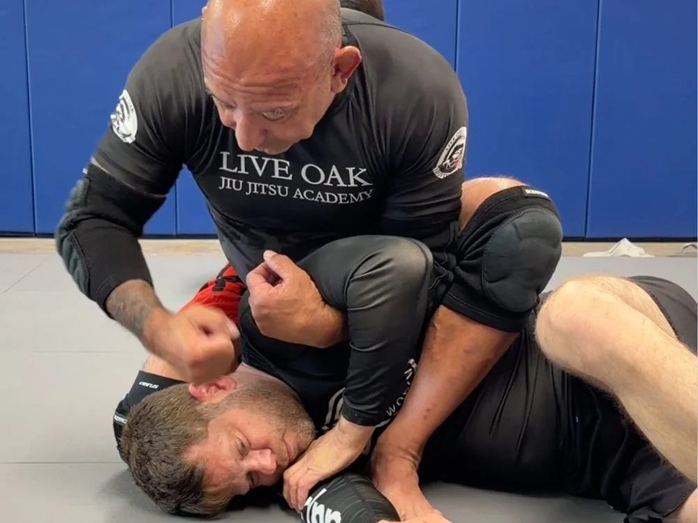 Two men practicing Brazilian Jiu-Jitsu on a mat, with one man on top in a controlling position and the other man on the ground beneath him, in a martial arts training facility with blue walls.