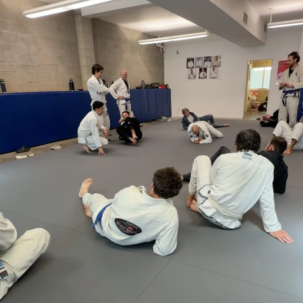 Brazilian Jiu-Jitsu students practicing techniques on the mats in a dojo with instructors and posters of fighters on the wall.