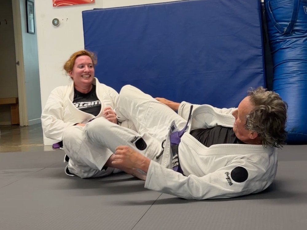 Two people in Brazilian Jiu-Jitsu gis on a mat, one woman smiling and the other man lying on the ground, practicing grappling techniques in a gym.