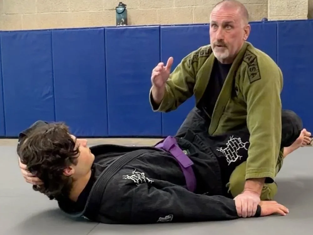 Martial arts training in a gym with two men, one in a black gi and purple belt on the ground, and the other in a green gi, kneeling and demonstrating a technique, with blue padding on the wall behind them.