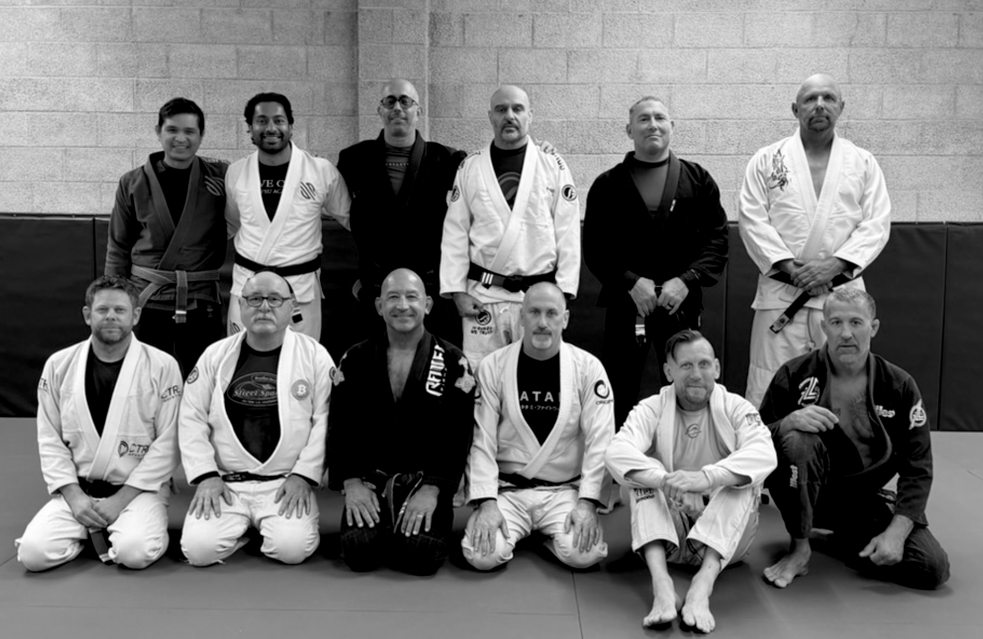 Group of Brazilian Jiu-Jitsu practitioners and instructors posing for a photo in a gym. They are dressed in gi uniforms, some with black belts, sitting and standing in front of a wall padded for training.
