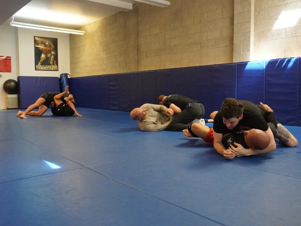 Two pairs of men practicing Brazilian Jiu-Jitsu on a blue mats in a gym, with one pair grappling and the other pair working on ground techniques.