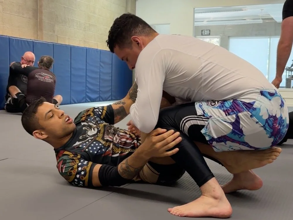 Two men practicing Brazilian Jiu-Jitsu on mats in a gym. One man is on his back, and the other is positioned over him, grabbing his collar and sleeve.