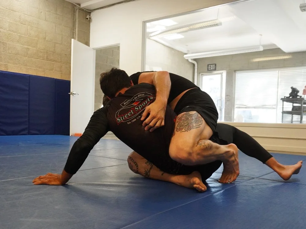 Two people practicing Brazilian Jiu-Jitsu on a blue mat, one in a black rash guard and the other in a black and red shirt, engaging in a grappling hold.