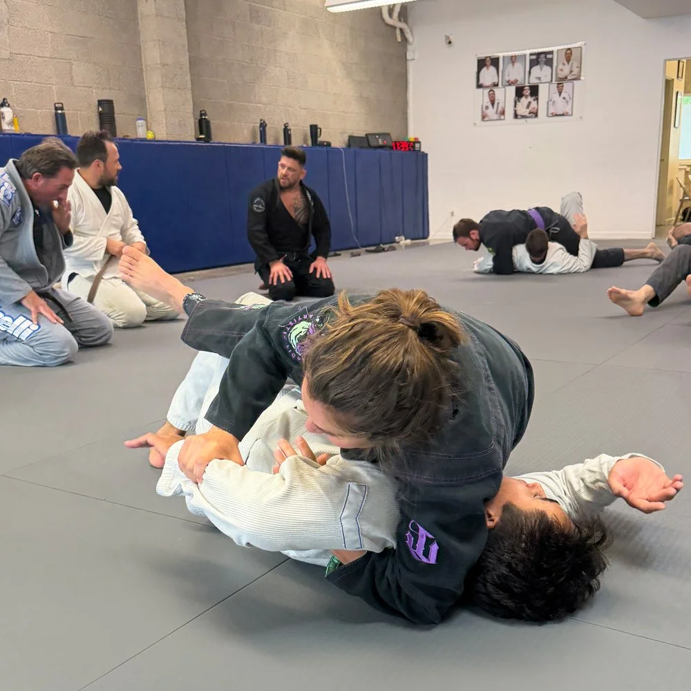 Martial arts class with students practicing Brazilian Jiu-Jitsu on mats, instructor observing, and photos on the wall.