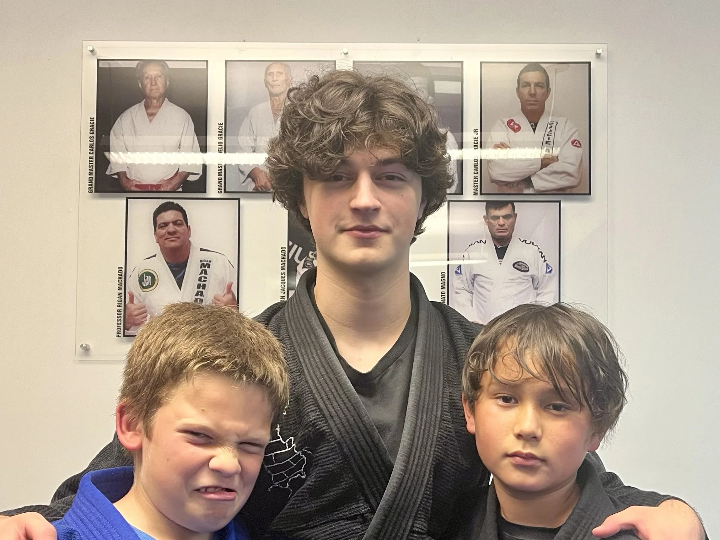 A young man with curly hair and a smile standing in front of a wall with framed pictures of martial arts practitioners and instructors.