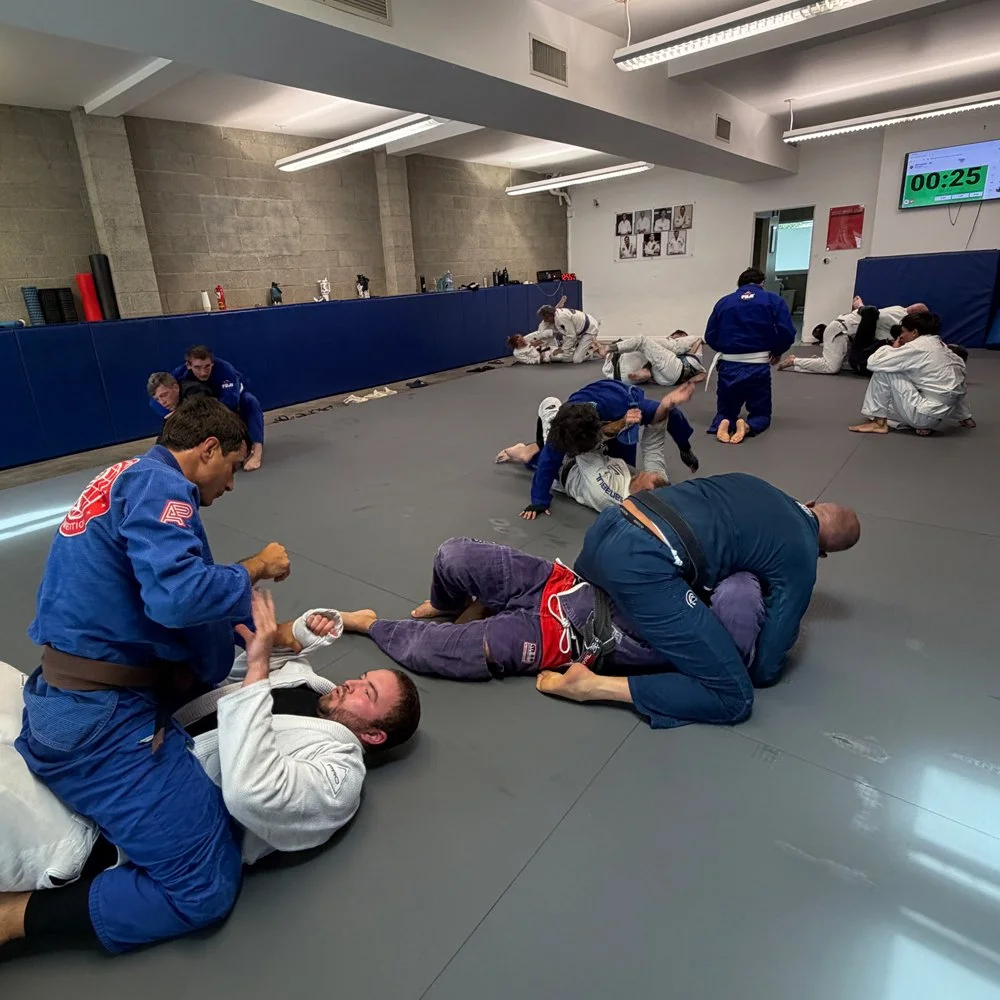 Brazilian Jiu-Jitsu students practicing techniques on mats in a training facility, with some students applying chokeholds and others practicing escapes under the supervision of instructors.