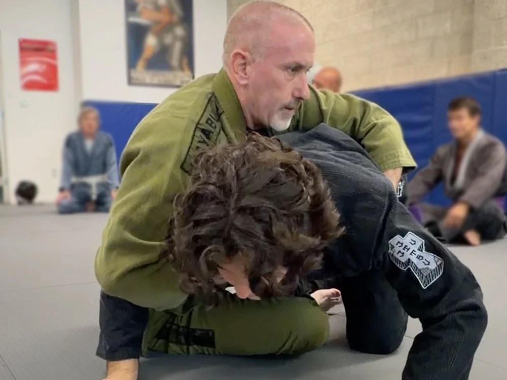 A martial arts class with a man in a green gi and a woman in a black gi practicing Brazilian Jiu-Jitsu techniques on a mat, with others observing in the background.