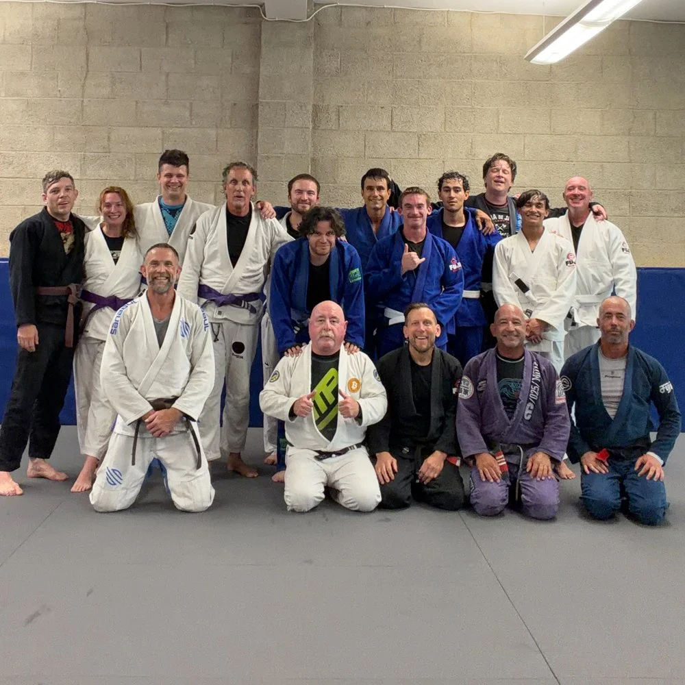Group of Brazilian Jiu-Jitsu practitioners in gis and rash guards, posing on mats in a training facility.