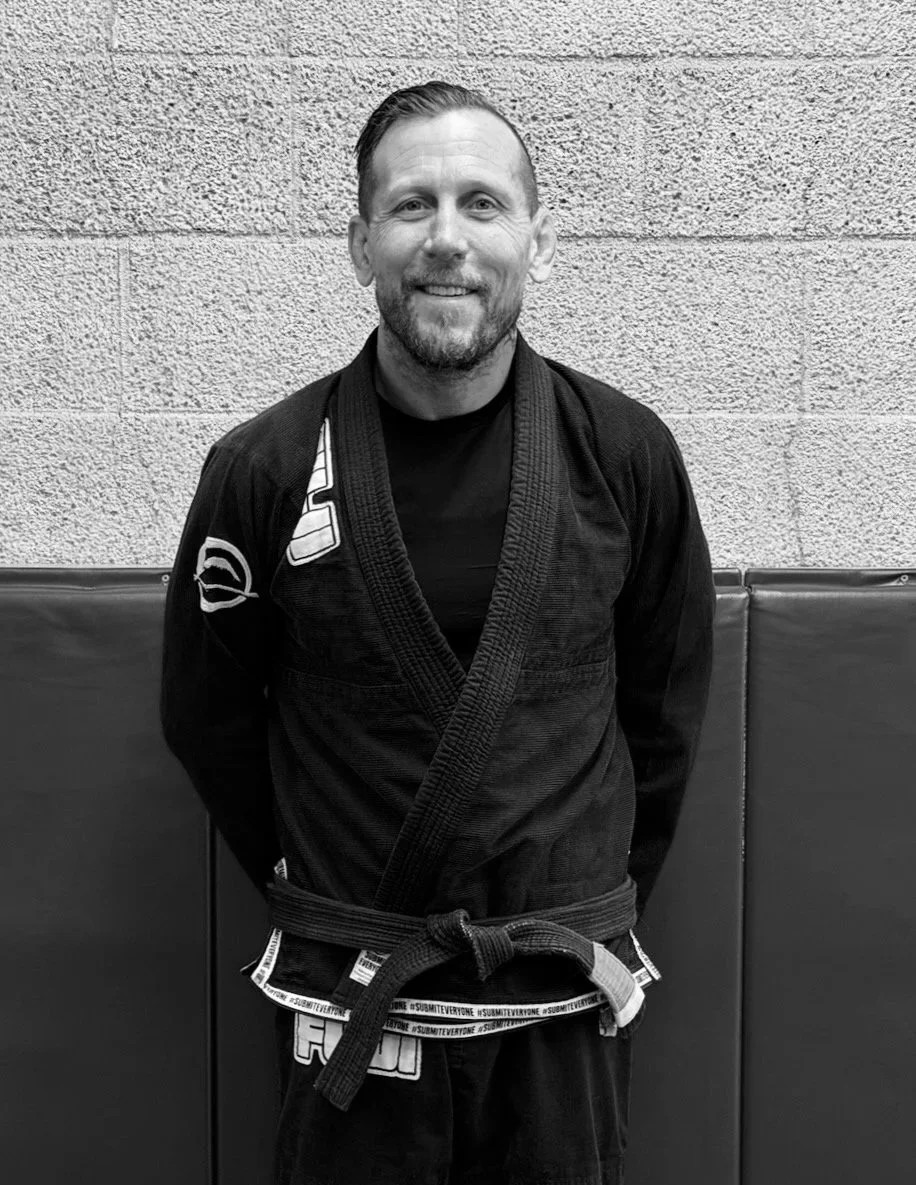 A man smiling, standing in a Brazilian Jiu-Jitsu gi, in front of a textured wall.