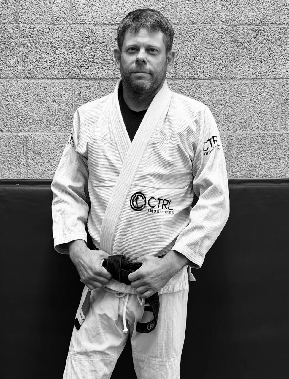 A man in a white Brazilian Jiu-Jitsu gi with a black belt standing against a textured wall.