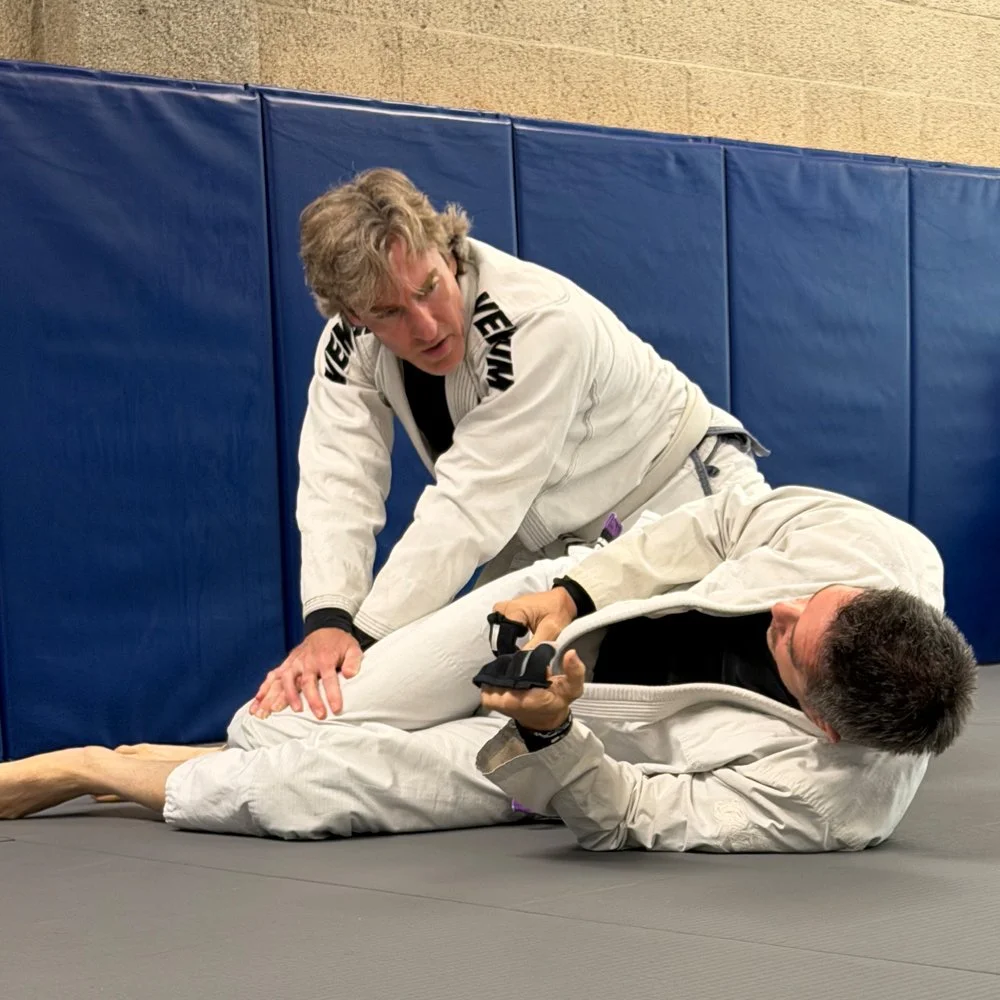 Two men practicing Brazilian Jiu-Jitsu on a blue mat in a training facility, with one man on top and the other on the ground holding a training gun while the man on top applies a choke.