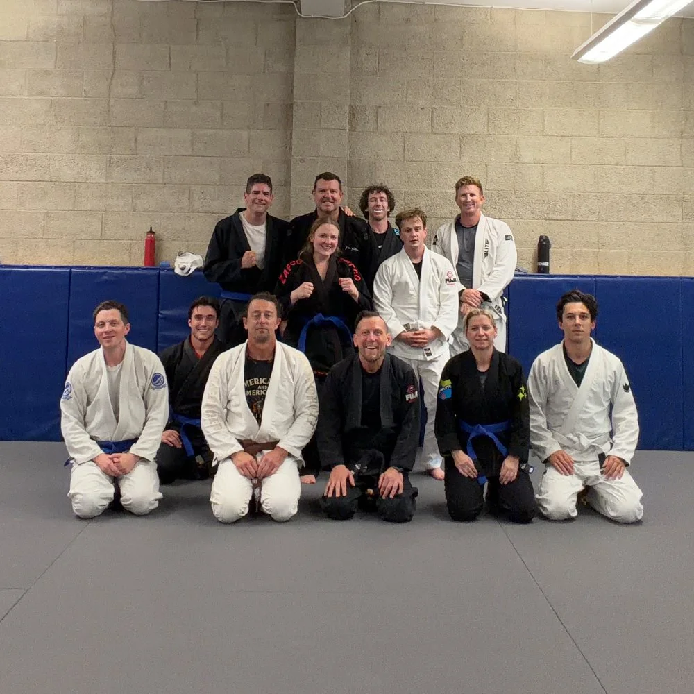 Group of people posing for a photo in a martial arts gym, some wearing white gis and others in black or hoodie, with blue belts, in front of a beige wall with blue padded mats.