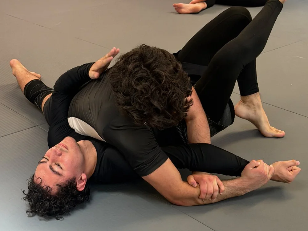 Two people practicing Brazilian Jiu-Jitsu on a gray mat, with one person on the ground in a black rash guard and the other person on top in a black shirt and black leggings, both focused on a grappling hold.