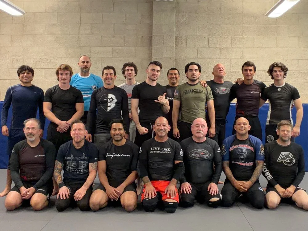 Group of men in MMA training gym, some wearing rash guards, sitting and standing on training mats, posing for photo.