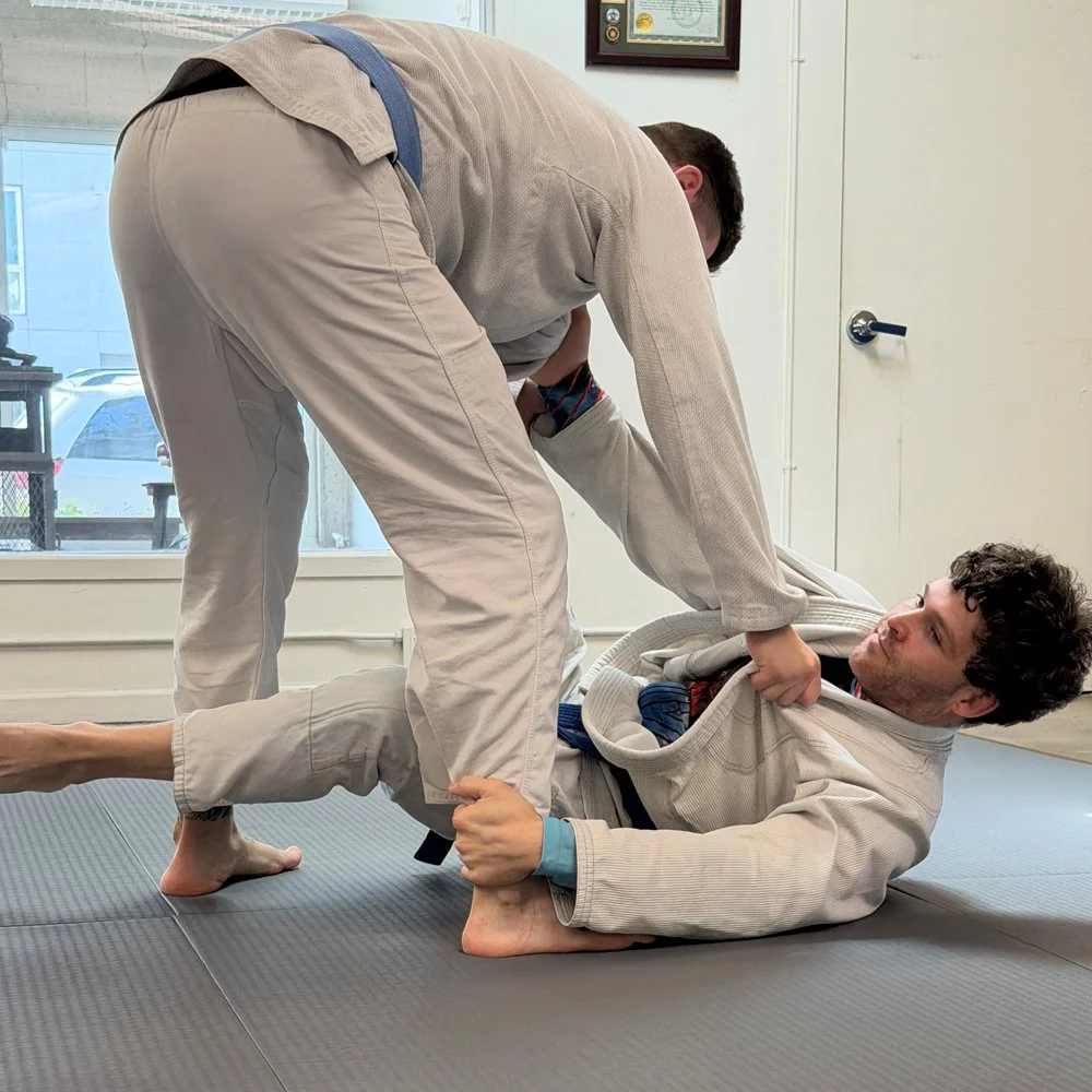 Two men practicing Brazilian Jiu-Jitsu, with one applying a guard position on the other on a mat in a training room.