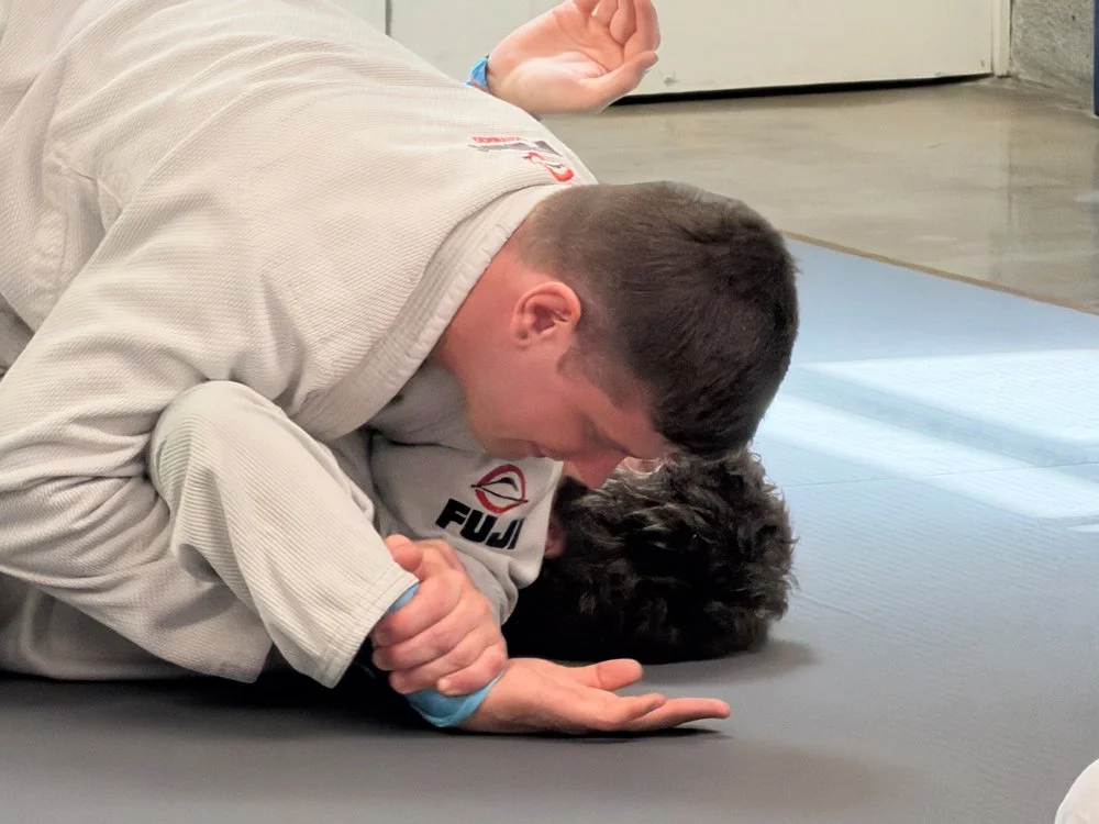 A person practicing Brazilian Jiu-Jitsu on a black dog, both lying on a mat in a training facility.