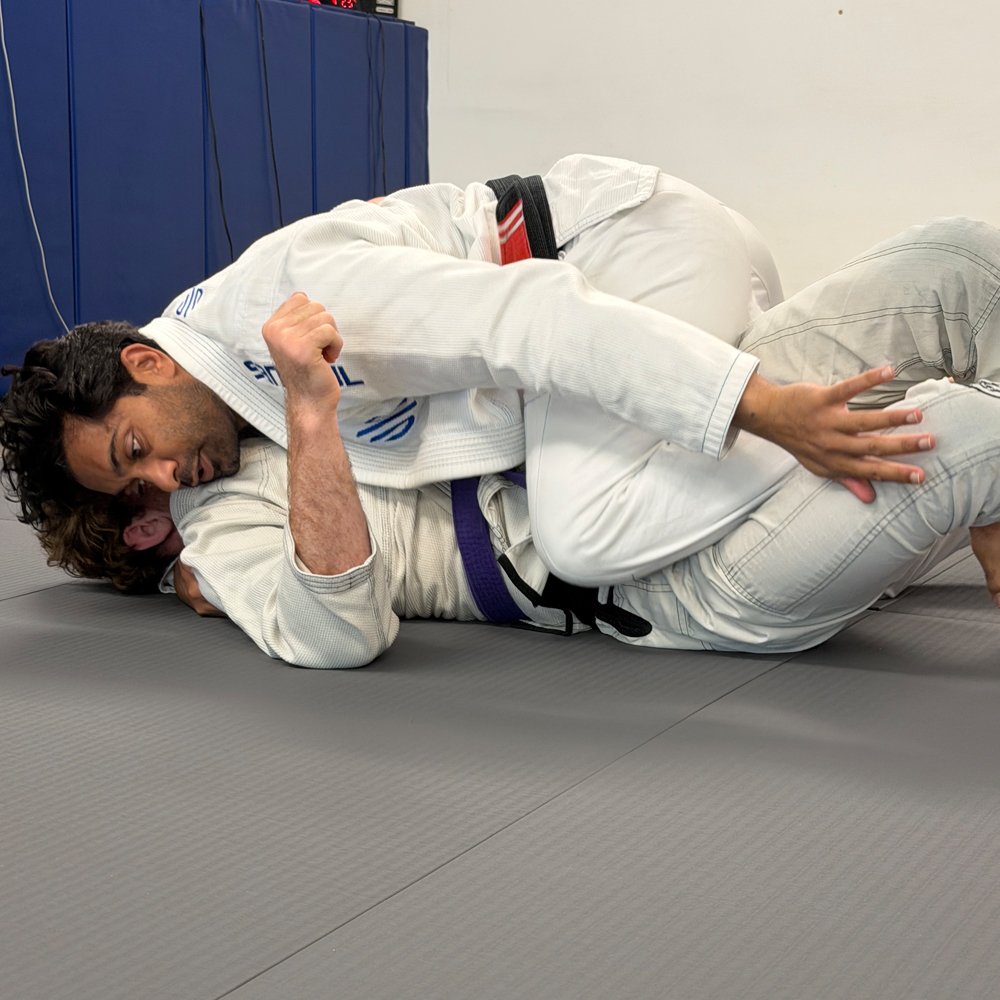 Two Brazilian Jiu-Jitsu practitioners in a grappling position on a gray mat, wearing white gi uniforms, with one on top of the other and both focused on their hold.
