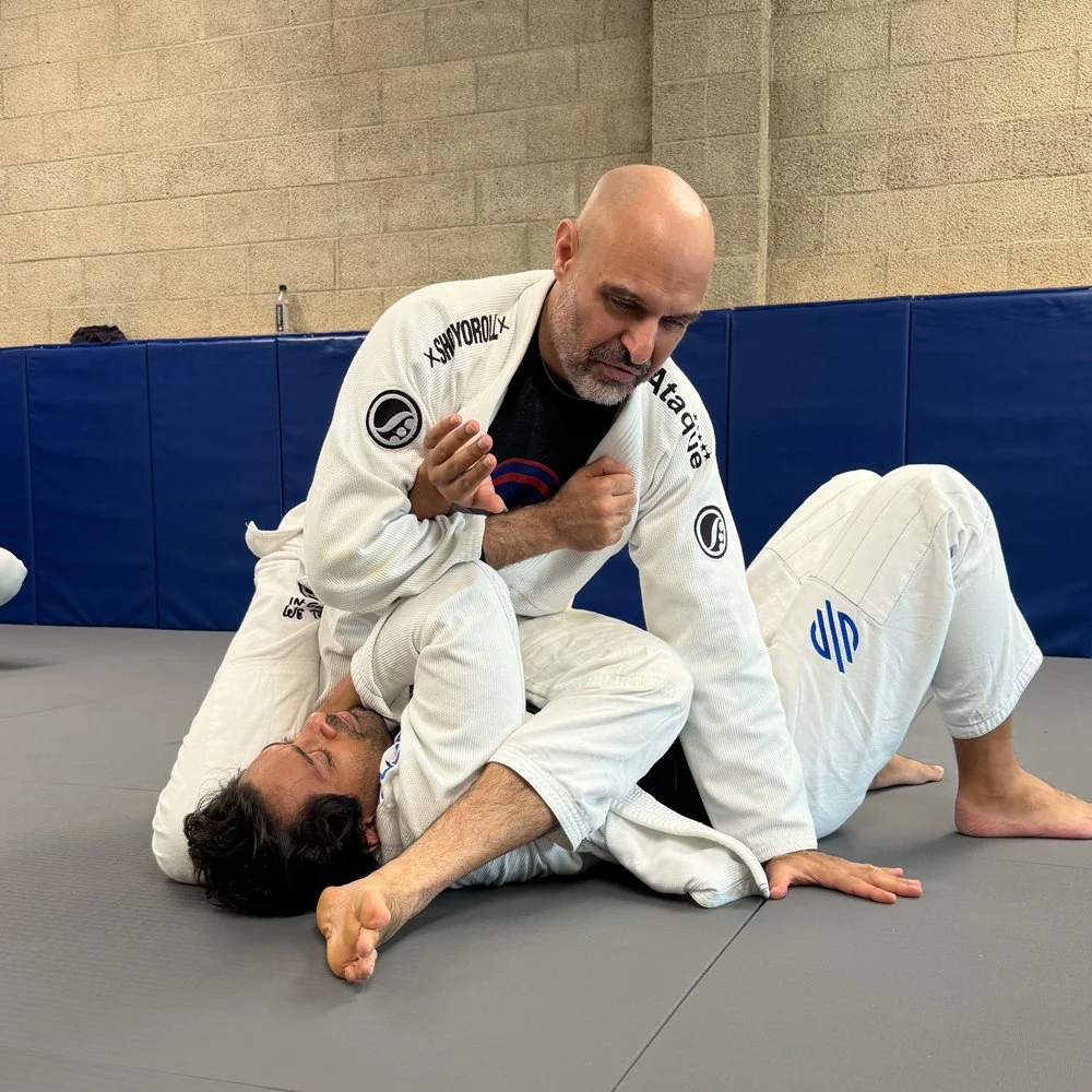 Two men practicing Brazilian Jiu-Jitsu on a mat indoors. One is applying a choke hold on the other who is lying on the ground.