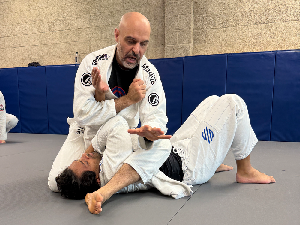 Two men practicing Brazilian Jiu-Jitsu on mats in a gym; one man is executing a technique while the other is on the ground, wearing gi uniforms.