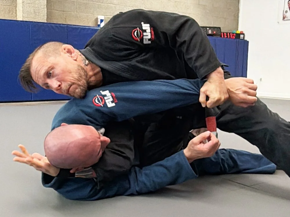 Two men practicing Brazilian Jiu-Jitsu, one in a black gi and the other in a navy gi, on a gray mat in a gym.