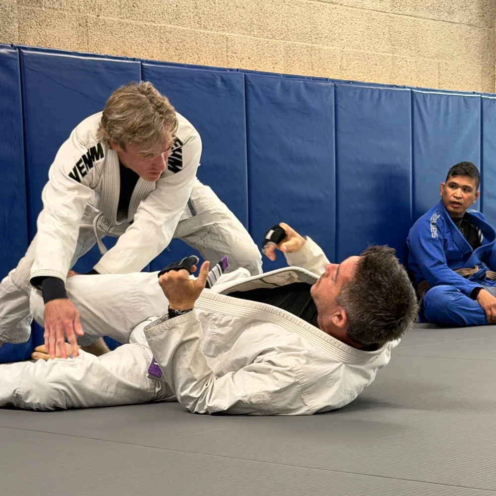 Two men practicing Brazilian Jiu-Jitsu on a mat, with one using a choke hold on the other. A third person in a blue gi sits nearby, observing.