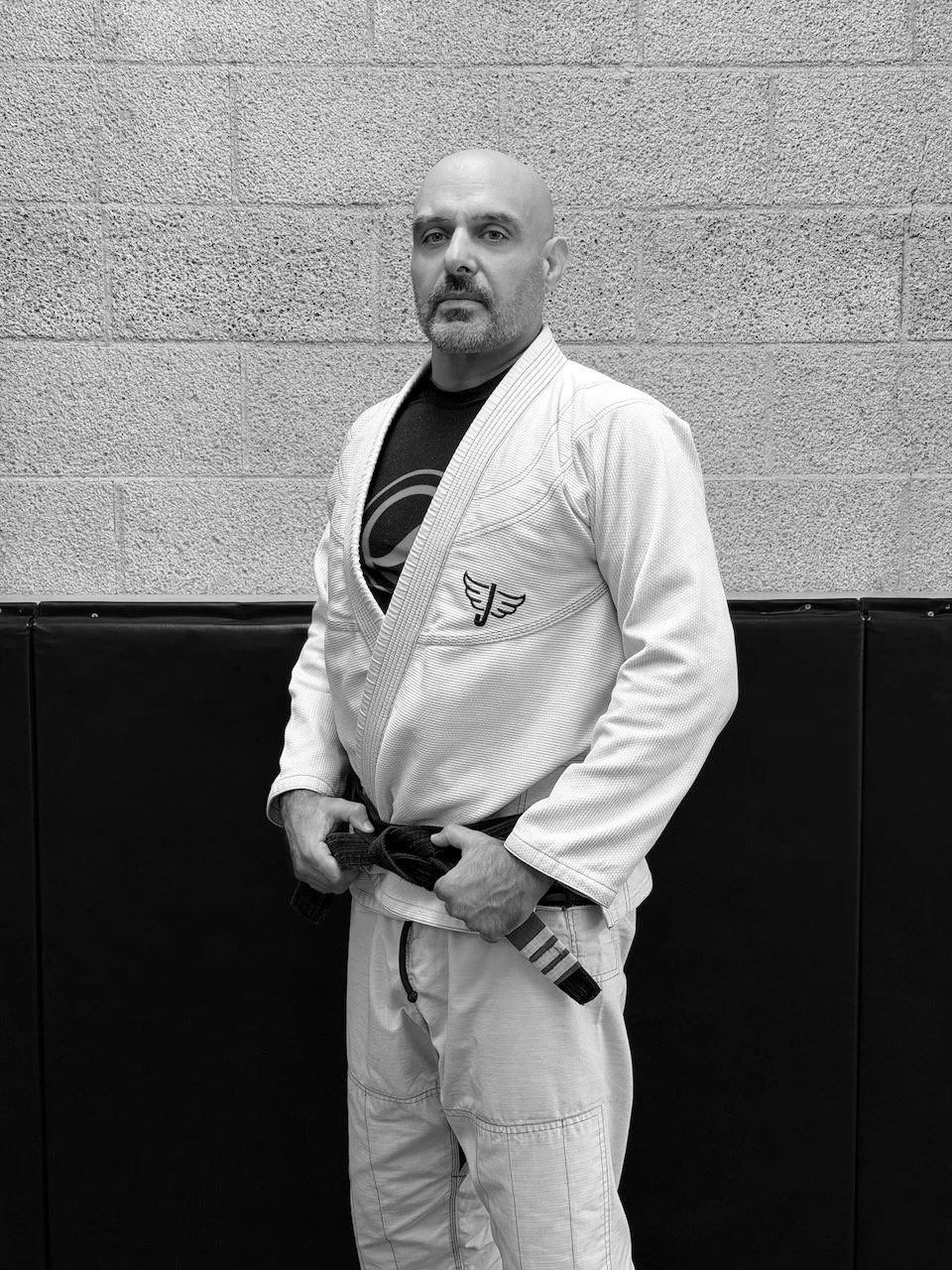 A man standing against a concrete wall, wearing a Brazilian Jiu-Jitsu gi and holding a black belt, with a serious expression.