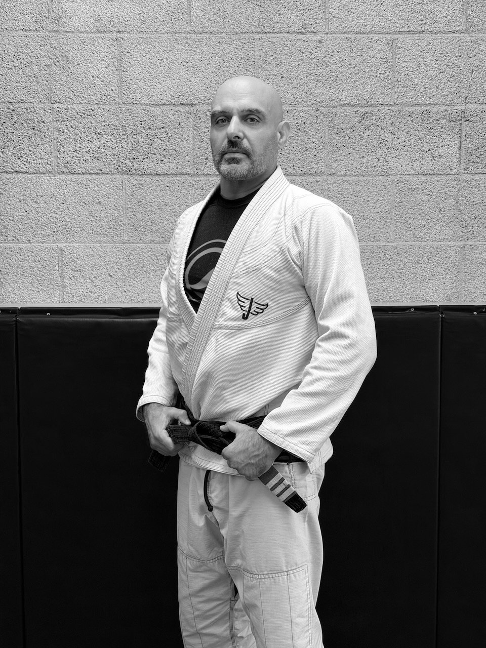 A man in a Brazilian Jiu-Jitsu gi holding a black belt, standing against a textured wall.