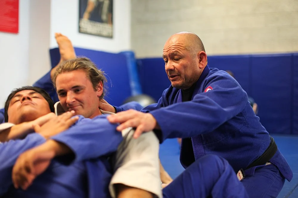 Martial arts class with two students practicing Brazilian Jiu-Jitsu, instructor demonstrating technique on two students in blue gis on padded mat