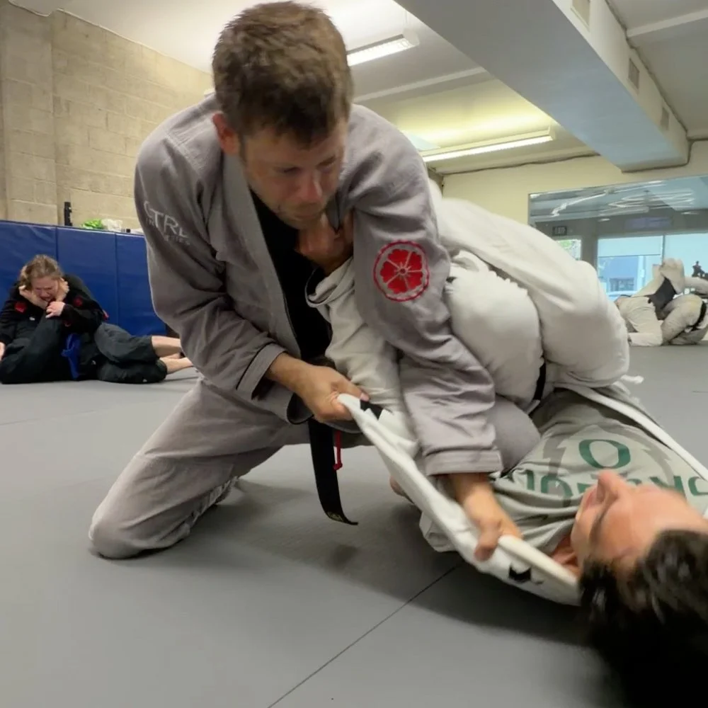 Two people practicing Brazilian Jiu-Jitsu, with one person in a grey gi applying a chokehold on another person in a white gi on a padded mat in a martial arts gym.